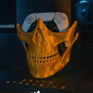 May include: A gold-colored skull-shaped controller holder with a white and black game controller inside. The base has the words "DEATH STRANDING" and the "PrintBROS" logo.