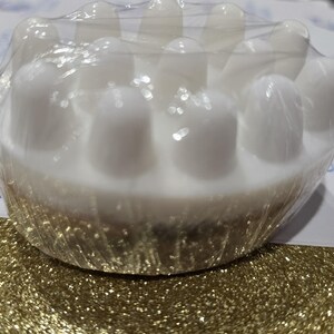 May include: A white, circular massage tool with rounded, raised nodules. The tool is wrapped in clear plastic and sits on a gold glitter surface. The design suggests a focus on relaxation and wellness.