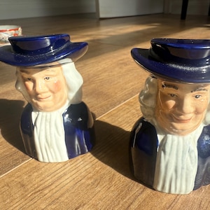 Op de afbeelding: Twee keramische beeldjes die een lachende man afbeelden met een blauwe hoed en jas en een witte cravat. De beeldjes hebben een vintage uitstraling en staan op een houten oppervlak. Een theekopje is zichtbaar op de achtergrond.