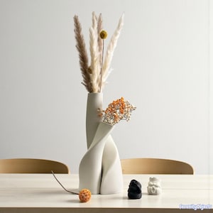 Op de afbeelding: Een moderne, gebroken witte keramische vaas met een gedraaid ontwerp, met gedroogd pampasgras en oranje bloemen. Een klein, rond, oranje decoratief object en twee kleine sculpturen staan op de tafel.