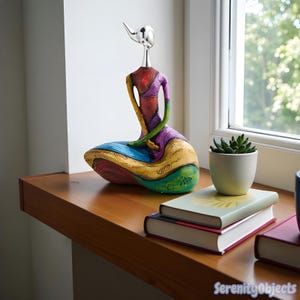May include: A vibrant, abstract sculpture of a seated figure with a silver head and neck. The figure is painted in bright colours including red, purple, blue, yellow, and green. The sculpture is displayed on a wooden surface near a window.