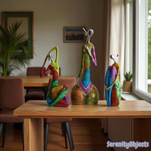 Könnte beinhalten: Drei farbenfrohe abstrakte Skulpturen auf einem Holztisch. Die Skulpturen zeigen stilisierte menschliche Figuren mit lebendigen Mustern und metallischen Akzenten. Die Figuren befinden sich in verschiedenen Posen, mit einer Mischung aus Grün-, Rot-, Blau- und Gelbtönen.