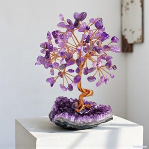 Könnte beinhalten: Dekorative Amethyst-Baumskulptur. Der Baum hat einen Stamm und Äste aus Kupferdraht, verziert mit Amethyst-Edelsteinblättern. Der Baum steht auf einem Sockel aus rohem Amethystkristall, was ein einzigartiges Aussehen erzeugt.