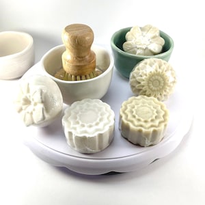 May include: A selection of handmade soaps with floral patterns, a wooden-handled brush, and small ceramic bowls. The soaps are white and light green. The brush is in a white bowl. The arrangement is on a white surface.