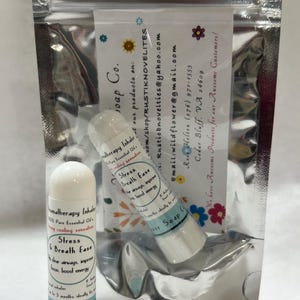 Puede incluir: Dos inhaladores de aromaterapia, etiquetados como "Stress & Breath Ease" y "Flower Soap", se muestran en una bolsa transparente sellada. Los inhaladores son blancos con texto azul y negro. También hay una tarjeta de visita.