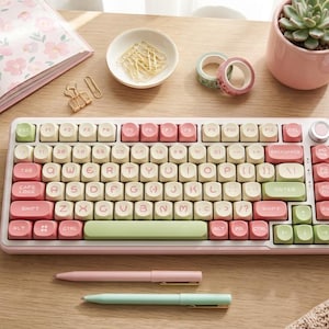 May include: A pastel-colored mechanical keyboard with pink and green keys sits on a wooden desk, surrounded by stationery. A floral-patterned notebook, pens, and a mug of coffee complete the aesthetic. The keyboard has a retro design.