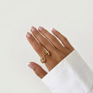 May include: A gold-toned ring featuring an open, fluid design with a central Arabic calligraphic element. The ring has a polished metallic finish and is worn on a finger, showcasing a curved, abstract shape that wraps around the digit.