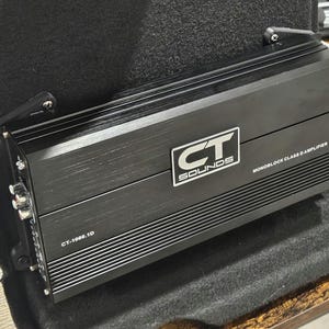 Peut inclure: Amplificateur Monobloc Classe D CT Sounds noir. L'amplificateur porte le texte "CT SOUNDS" dans un rectangle blanc et le numéro de modèle "CT-1000.1D". L'amplificateur est monté dans un véhicule.