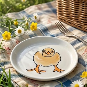 May include: A white ceramic plate with a cartoon chick illustration. The chick is yellow with black outlines and orange feet. The plate is on a blue and white checkered picnic blanket, with a wicker basket and fork in the background.