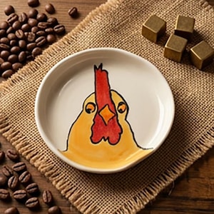 May include: A small, round, white ceramic dish with a hand-painted yellow and red rooster design. The dish sits on a burlap cloth, surrounded by coffee beans and small gold-colored cubes.