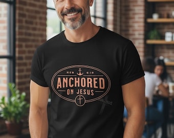 May include: Black t-shirt with the text "ANCHORED ON JESUS" in a peach-colored oval design. The shirt also features an anchor and cross symbol. The man in the image is wearing blue jeans.