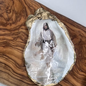 Puede incluir: Una obra de arte en concha de ostra con borde dorado, que muestra una imagen de Jesús. La concha descansa sobre una superficie de madera. La imagen de Jesús está en tonos suaves y neutros.