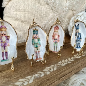 Peut inclure: Quatre plats décoratifs en coquilles d'huîtres, chacun avec une figurine de soldat casse-noisette miniature. Les coquilles ont des bords peints en or et sont présentées sur de petits supports dorés. Les casse-noisettes sont de couleurs variées.
