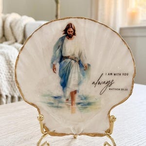 May include: A white scallop shell with gold-painted edges, displayed on a gold metal stand. The shell features a printed image of a figure in blue and white robes walking on water, with the text "I AM WITH YOU always MATTHEW 28:20" printed in black.
