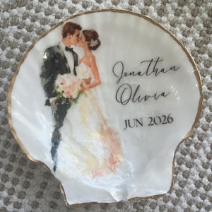 May include: White seashell-shaped dish with a gold rim. The dish features a watercolor image of a kissing couple, with the names "Jonathan Olivia" and the date "JUN 2026" in black script. The dish is on a textured surface.