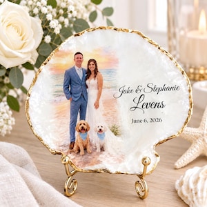 Puede incluir: Una placa decorativa blanca con forma de concha y borde dorado sobre un soporte de metal. Incluye una fotografía impresa de una pareja y dos perros en la playa, junto con el texto Jake & Stephanie Levens y June 6, 2026.