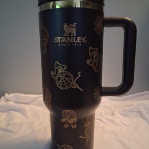 Puede incluir: Taza de viaje Stanley negra con asa, con gr&aacute;ficos dorados de Spider-Man. La taza tiene una tapa negra y un borde dorado. El logotipo de Stanley y "SINCE 1913" est&aacute;n impresos en dorado.