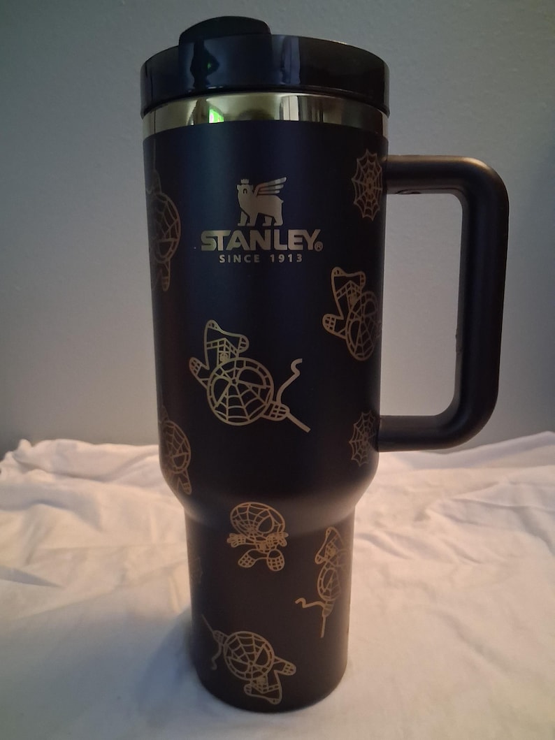 Puede incluir: Taza de viaje Stanley negra con asa, con gr&aacute;ficos dorados de Spider-Man. La taza tiene una tapa negra y un borde dorado. El logotipo de Stanley y "SINCE 1913" est&aacute;n impresos en dorado.