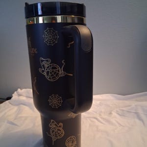 Puede incluir: Vaso t&eacute;rmico negro con asa, con dise&ntilde;os dorados de telara&ntilde;as y brujas. El vaso tiene una tapa negra y una banda dorada. El logotipo de Stanley es visible en el lateral.