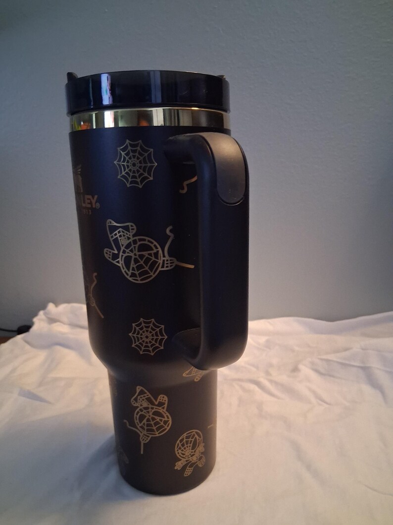 Puede incluir: Vaso t&eacute;rmico negro con asa, con dise&ntilde;os dorados de telara&ntilde;as y brujas. El vaso tiene una tapa negra y una banda dorada. El logotipo de Stanley es visible en el lateral.