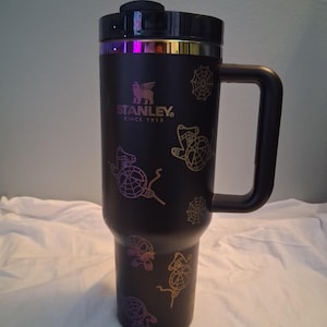 K&ouml;nnte beinhalten: Schwarzer Stanley-Reisebecher mit Griff und Deckel. Der Becher hat ein Design aus Spinnen und Spinnweben in Lila-, Gelb- und Oranget&ouml;nen. Der Text "STANLEY SINCE 1913" ist auf dem Becher aufgedruckt.