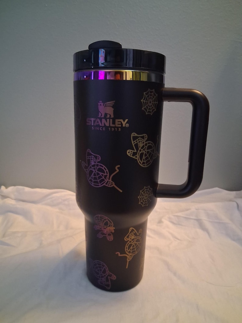 K&ouml;nnte beinhalten: Schwarzer Stanley-Reisebecher mit Griff und Deckel. Der Becher hat ein Design aus Spinnen und Spinnweben in Lila-, Gelb- und Oranget&ouml;nen. Der Text "STANLEY SINCE 1913" ist auf dem Becher aufgedruckt.