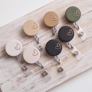 May include: A collection of seven circular badge reels in various matte colors including white, beige, tan, olive green, and black. Each badge reel features a small, gold-toned outline of a heart on the surface and a clear plastic strap with a metal snap.