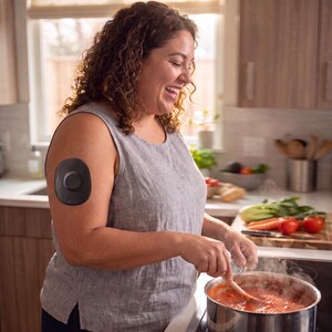 Puede incluir: Un sensor de glucosa continuo de forma ovalada y color gris oscuro est&aacute; adherido a la parte superior del brazo. El dispositivo presenta un acabado liso y mate con un centro circular ligeramente elevado, ofreciendo un aspecto compacto y discreto.