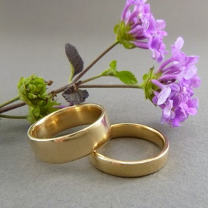 May include: Two gold wedding bands, one slightly larger than the other, are lying on a gray surface. A sprig of purple flowers with green leaves is behind the rings.