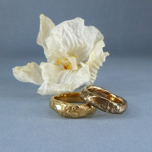 May include: Two gold wedding bands with a textured finish, resting on a blue background with a white flower.