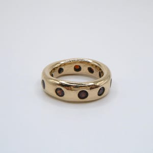 Può includere: Un anello a fascia color oro caratterizzato da una serie di pietre preziose circolari rosso scuro, distanziate uniformemente lungo l'intera circonferenza esterna della superficie metallica lucida, creando un motivo continuo di pietre.