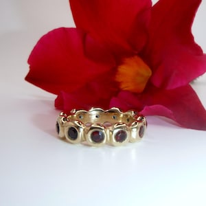 May include: A gold ring featuring a sequence of round, dark red gemstones set in individual bezels. The ring is set against a white backdrop, with a large, vibrant red flower in the background.