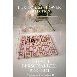 Pode incluir: Uma vitrine de acrílico transparente com chocolates personalizados para um chá de bebê. Cada quadrado de chocolate apresenta um desenho de borboleta e o nome "Alya Lina". A vitrine é decorada com flores brancas e o texto "Luxury Baby Shower Collection".