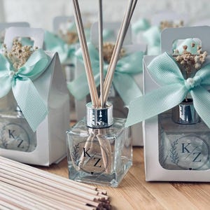 Könnte beinhalten: Ein quadratischer Glas-Diffusor mit silbernem Hals und Holzstäbchen, verziert mit einem kreisförmigen Logo mit den Buchstaben K/Z. Die Flasche wird in einer weißen Geschenkbox mit hellblauem Band und Trockenblumen sowie zusätzlichen Stäbchen präsentiert.