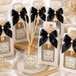 Op de afbeelding: Een collectie vierkante glazen rietdiffusers met gouden doppen en houten stokjes. Elke fles heeft een label met een bloemenkrans, de letters G|A en de datum 8.27.2026. De diffusers zijn verpakt in witte doosjes met zwarte satijnen strikken.