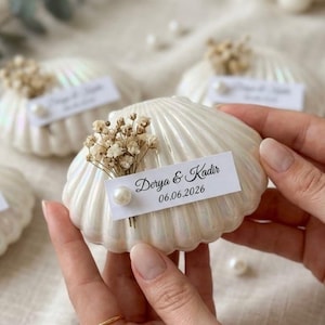 Puede incluir: Una concha marina blanca iridiscente decorada con un pequeño racimo de flores secas, una perla blanca y una etiqueta de papel rectangular con el texto Derya & Kadir y la fecha 06.06.2026 impresos en una fuente de estilo caligráfico negro.