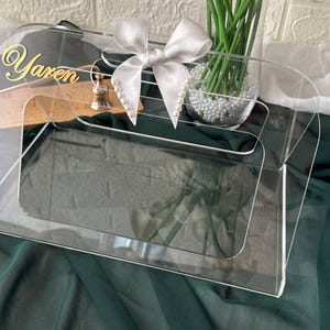 Pode incluir: Uma caixa retangular para lenços de papel em acrílico transparente com o nome Yaren em letras douradas no canto superior esquerdo. A caixa é decorada com um laço de cetim branco adornado com pequenas pérolas brancas nas pontas.