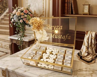 May include: A glass box with gold trim, filled with white candies and small chocolate bars. The box is labeled with the names "Beyza Mustafa" in gold lettering. A floral arrangement and a decorative scarf are also visible.