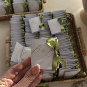 May include: A collection of small, white square cards tied with olive green satin ribbons. The cards have gold script lettering and the date 14.01.2024. The cards are arranged in a woven basket.