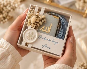 May include: A square gift box containing a small grey book with gold Arabic calligraphy, a blue beaded tasbih with a tassel, and a cluster of dried white flowers. A white wax seal with a leaf design sits on the box, which features the text Dualarla büyü Aras.