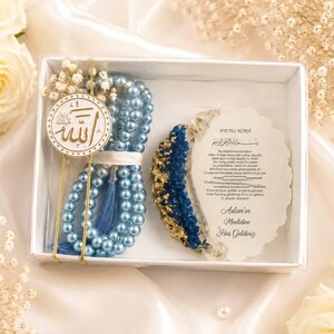 May include: A white gift box containing a string of light blue pearl prayer beads with a blue tassel, a circular white pendant with gold Arabic calligraphy, and a white resin plaque featuring blue and gold accents and printed Turkish text.