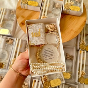 May include: A white gift box containing a small velvet-covered book with gold Arabic calligraphy, a glass perfume bottle with a wooden cap, a circular white resin coaster with gold edges and printed text, and a strand of white pearl prayer beads.
