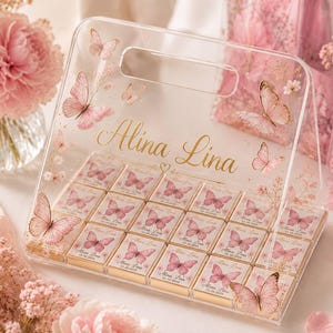 May include: A clear acrylic display stand featuring a handle and gold lettering that reads Alina Lina. The stand holds twenty small square chocolate bars, each wrapped in white paper with a pink butterfly graphic and the name Alina Lina.