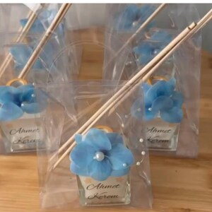 May include: Several small, clear glass bottles with blue floral decorations and wooden sticks are displayed in clear plastic bags. Each bottle has a gold-colored handle and the name "Ahmet Kerem" is printed on the front.