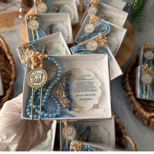 May include: Several gift boxes with blue beaded prayer beads, gold accents, and a decorative medallion with Arabic script. Each box contains a card with text. The boxes are arranged in a woven basket.