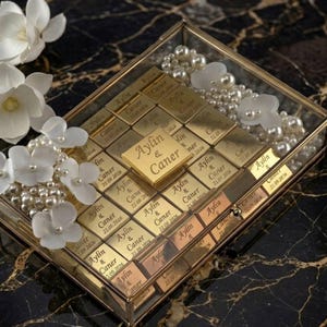 Puede incluir: Una caja de cristal con marco dorado, llena de barras doradas grabadas con nombres y fechas. Flores blancas y perlas adornan la caja. La caja descansa sobre una superficie de mármol negro y dorado. Los nombres 'Aylin & Caner' son visibles.