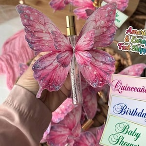 Puede incluir: Un frasco de perfume rosa con forma de mariposa y tapón dorado. Las alas de mariposa están decoradas con purpurina y un degradado de tonos rosas. El frasco tiene una etiqueta con el texto "Morelis RV 06-27-2026". Hay tres tarjetas con las palabras "Quinceañera", "Birthday" y "Baby Shower".
