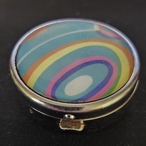 Puede incluir: Un pastillero pequeño y redondo con un marco de metal plateado. La tapa presenta un diseño abstracto colorido con círculos concéntricos en tonos de azul, amarillo, rosa y blanco. El pastillero está cerrado y sobre una superficie oscura.