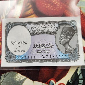 May include: A five-pound Egyptian banknote with Arabic script and a pharaoh's image. The note has a blue and purple floral design and a portrait on the right. It is encased in a clear plastic sleeve.