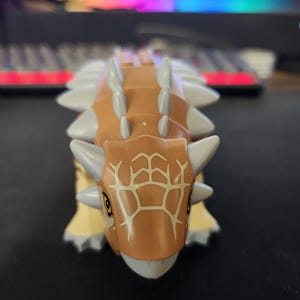Puede incluir: Un juguete de dinosaurio marrón y beige con púas grises y una cabeza estampada. El juguete tiene patas amarillas y ojos pequeños y amarillos. El dinosaurio es una figura de juguete.